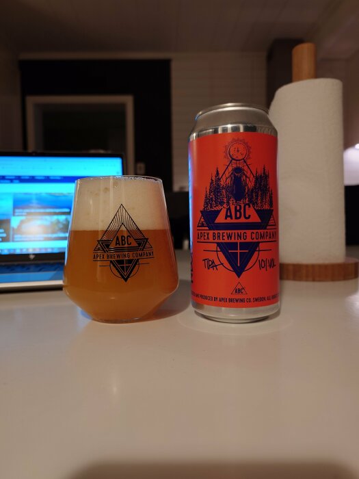 Ölglas med logga bredvid burk från Apex Brewing. Dator och pappershandduk i bakgrunden.