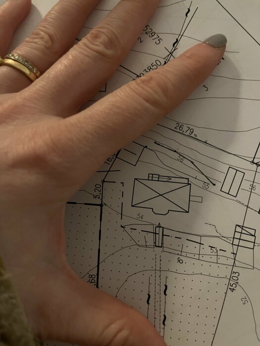 En hand med ring på teknisk ritning som visar linjer, mått och konstruktionsdetaljer.