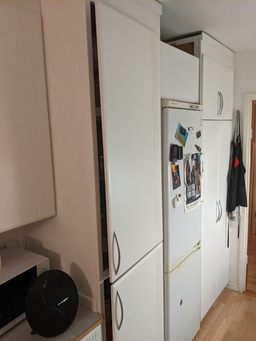 Kökshörn med vita skåp, kylskåp dekorerat med magneter, mikrovågsugn och robotdammsugare på golvet.
