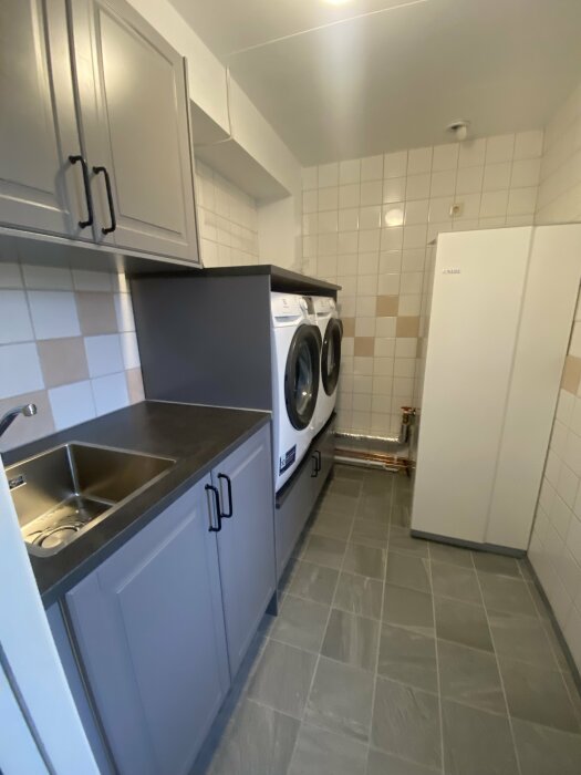 Kök med tvättmaskiner, skåp, diskho och kylskåp; modernt, kompakt, grått och vitt.
