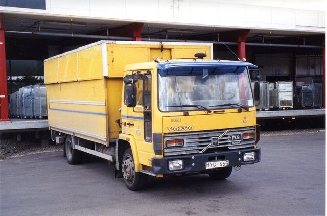 Gul Volvo FL6 lastbil framför en byggnad med lastkaj och fönster synliga.