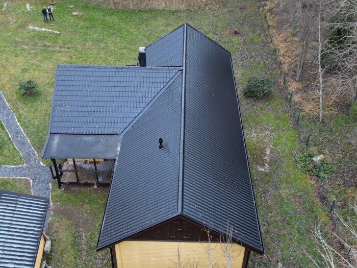 Flygbild av svart tak på två byggnader, personer i trädgården, omgivna av naturen.