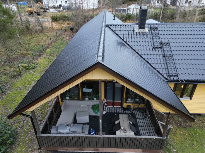 Ett tvåvåningshus med svart tak, terrass, utemöbler, omgiven av grönska. Vy uppifrån.