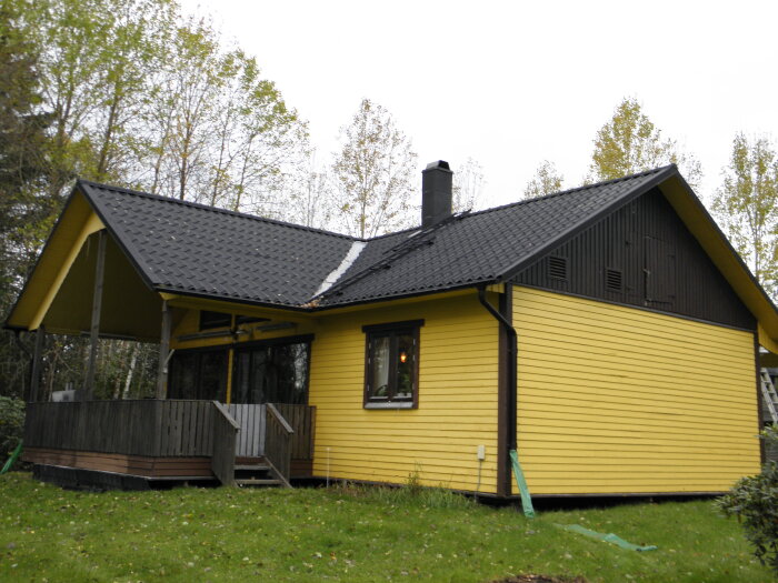 Gult hus med svart tak, skorsten och veranda omgiven av grönska.