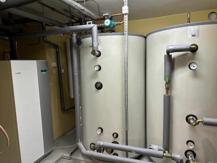 Värmepump och varmvattenberedare i ett teknikrum med rör och ledningar.