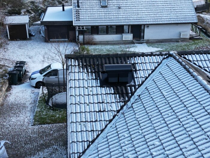 Snödäckade tak och uppfart vid ett bostadshus med parkerade bilar och soptunnor.