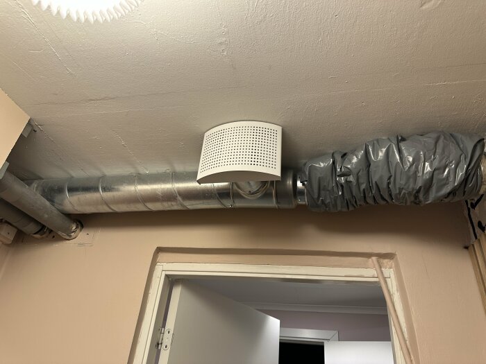 Ventilation i taket med isolerad rörledning och vit ventilationsgrill över en dörröppning.