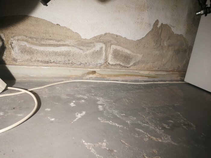 Fukt- och mögelskador längs en betongvägg, vit kabel på golv, tecken på vattenskador.