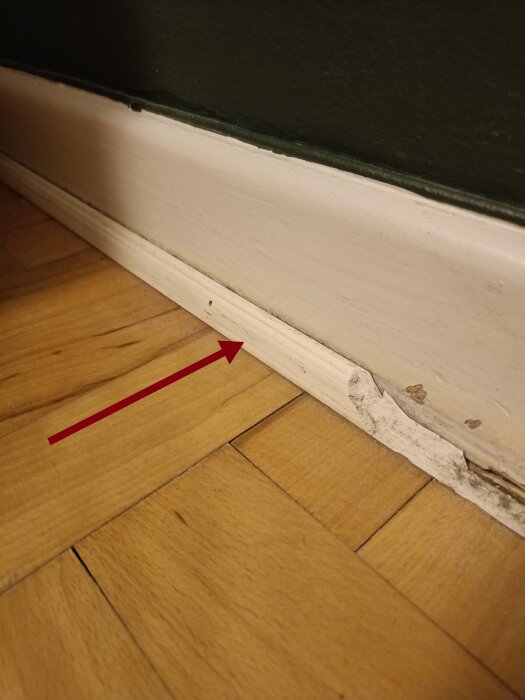 Skadad list vid golv och vägg, träparkett, röd pil pekar på skadan.