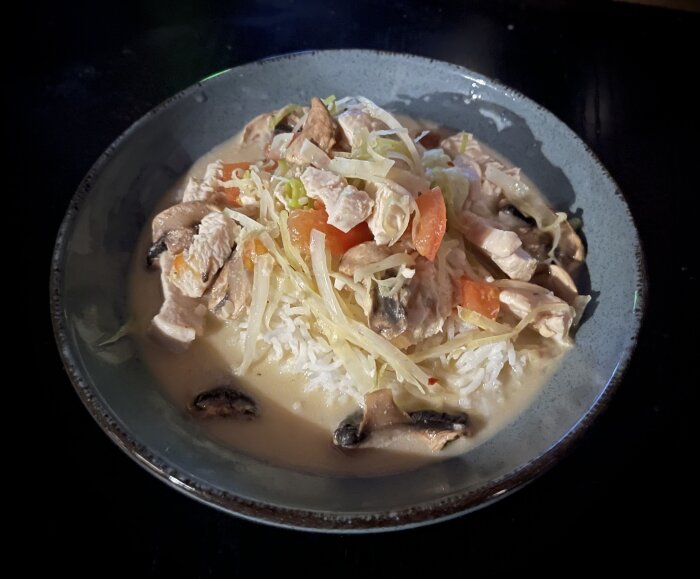 En skål med gräddig kycklingpasta toppad med riven ost och grönsaker på en mörk bakgrund.