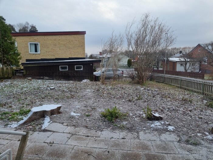 Trädgård med lätt snötäcke, gult tegelhus, träskjul, staket, blottade buskar, molnig himmel, vinter eller tidig vår.