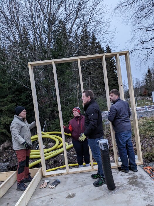 Fyra personer diskuterar byggprojekt av träram utomhus i dagsljus. Verktyg och byggmaterial synliga.