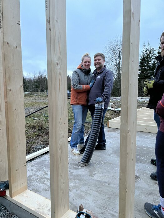 Två leende personer kramas på en konstruktionsplats med trästomme och byggmaterial runt omkring.