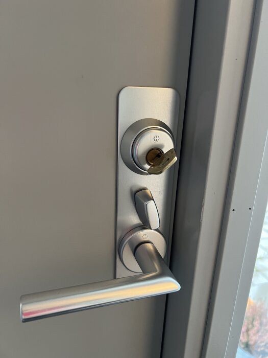 Dörrhandtag i metall med nyckel i låscylinder vid grå dörr.