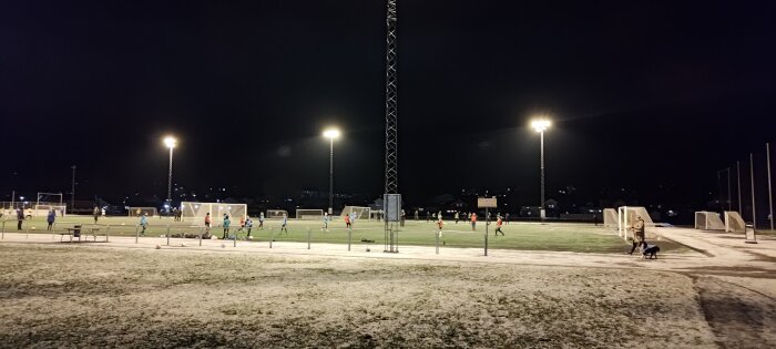 Kvällsträning på snötäckt fotbollsplan med konstgräs under lysande strålkastare, person promenerar med hund.