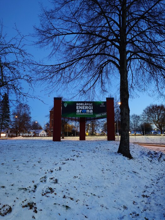 Skymning, snötäckt mark, trädstam, bar grenverk, reklamskylt, "BORLÄNGE ENERGI", gatlyktor, klart himmel.