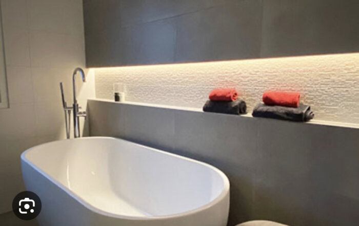 Modernt badrum med fristående badkar, gråa väggar, indirekt belysning och röda handdukar.