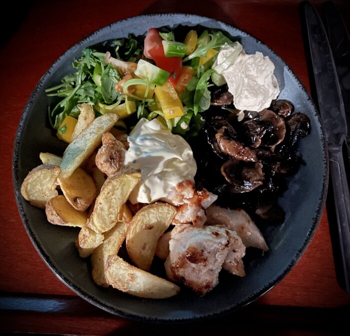 Maträtt med grillad kyckling, stekt potatis, sallad, svamp, och gräddfil serveras på svart tallrik.