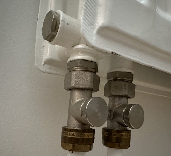 Radiatorventiler och röranslutningar, metall och vit, mot en vägg.