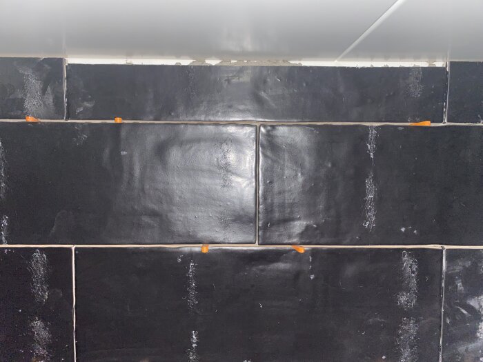 Svarta kakelplattor med vita fogar, ojämn yta, och återstående byggsmuts. Ljusrör ovanför belyser väggen.
