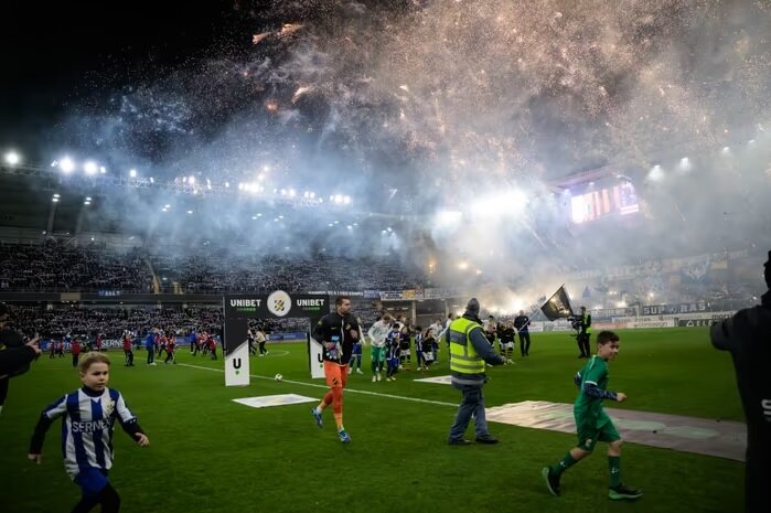 Fotbollsmatch med fyrverkerier, rök, fotbollsspelare och barn på en belyst arena vid kvällstid.