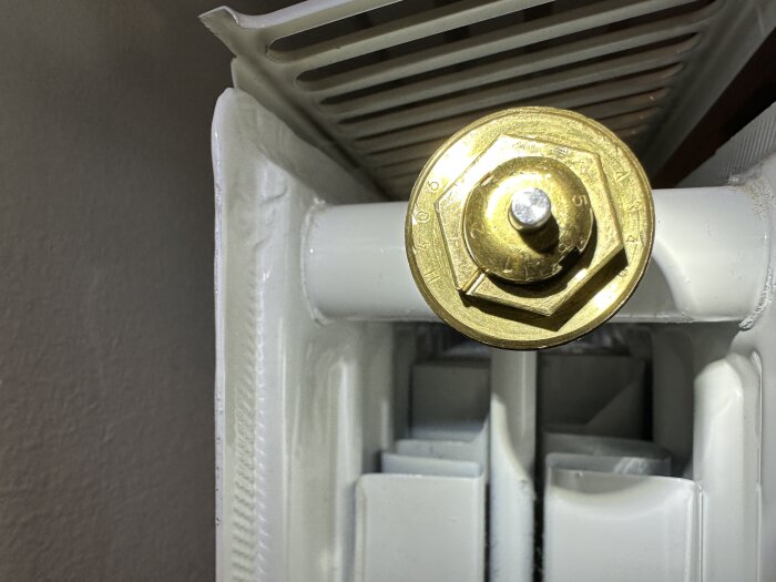 Radiatortermostat med guldfärgad ratt, vita rör och del av vit radiator syns.