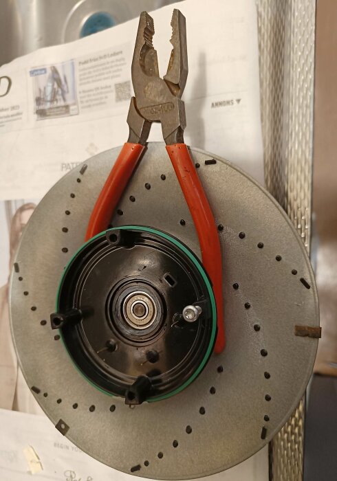 En tång hängande på en cirkulär metalldel med perforering och centrala komponenter, placerad framför en dokumentväska.