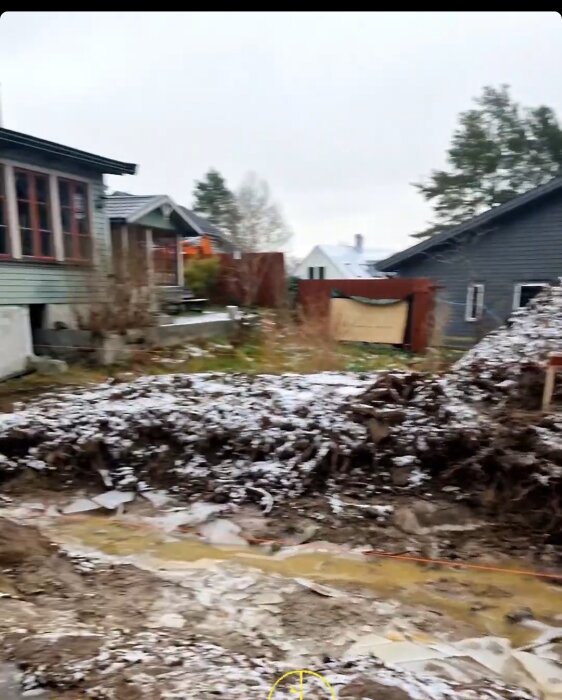 Otydligt, grumligt foto av en byggarbetsplats med snötäckta högar av jord, materialrester, och hus i bakgrunden.