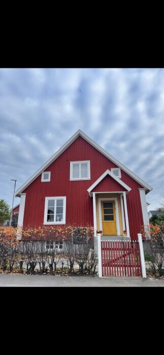 Ett rött trähus med vita knutar, gult dörröverstycke, staket och molnig himmel.