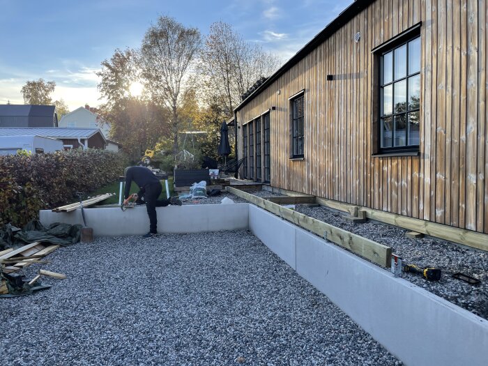 Person arbetar utomhus vid träbyggnad med verktyg, grus och byggmaterial på hösten.