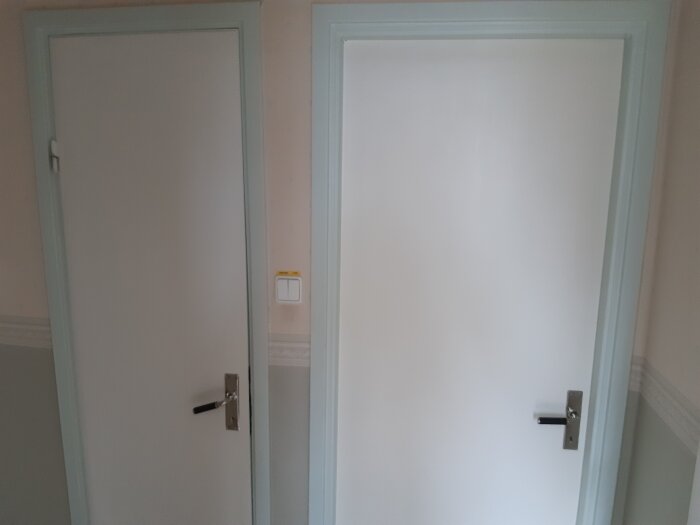 Två vita dörrar i en vägg, en öppen och en stängd, med metallhandtag och ljust rum.