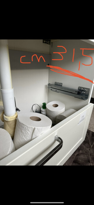 Inredningsskåp med rör, toalettpappersrullar och skena, märkt med röd text och linje.