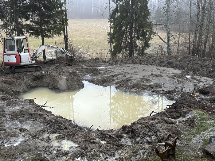 Grävmaskin vid lerig utgrävning med vattenansamling, skogsbakgrund, disigt väder.