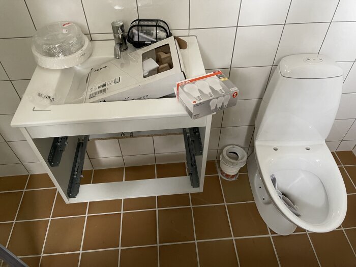 Badrum under renovering med wc-stol, demonterad tvättställsenhet, verktyg och förpackningar.