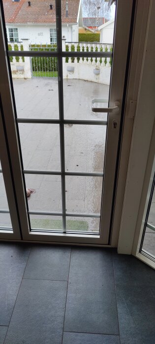 Glaserad dörr med utsikt över våt uteplats, vit vägg, grönt staket, krukor. Inomhus grått kaklat golv.