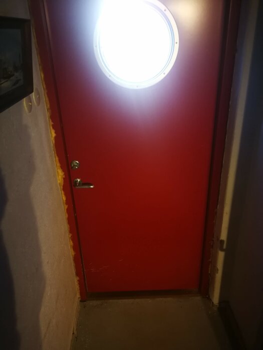 Röd dörr med rund ljuskälla ovanför, dörrhandtag till höger, väggar behöver målning eller reparation.