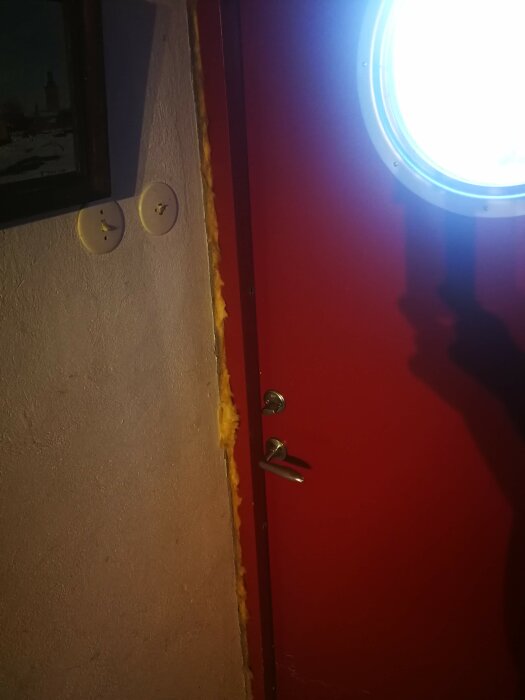 Röd dörr med handtag, cirkelformat ljus, skugga, dubbla vägguttag, isoleringsskum vid karm.