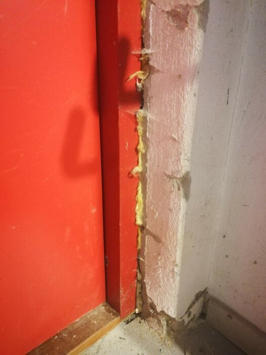 Röd dörrkarm med gul isolering och vitvägg, ofullständig finish, byggämnen synliga, behov av reparation.