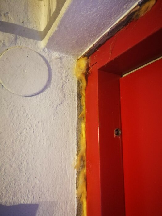 Röd dörrkarm med synlig gul isolering och vit vägg, tecken på slitage eller skada.
