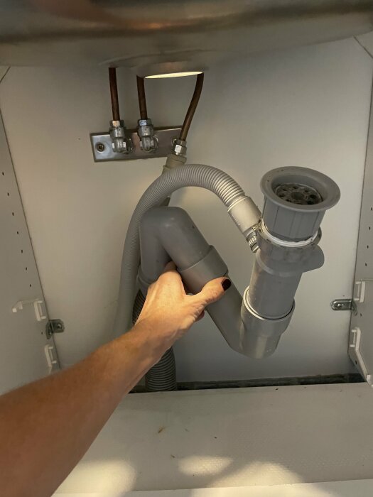 En hand som justerar avloppsröret under en diskbänk med synliga vattenledningar.