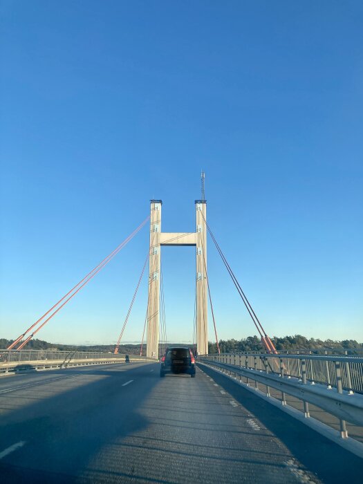 Hängbro med röda kablar, bil på vägen, klarblå himmel utan moln, dagtid, skuggor på asfalten.