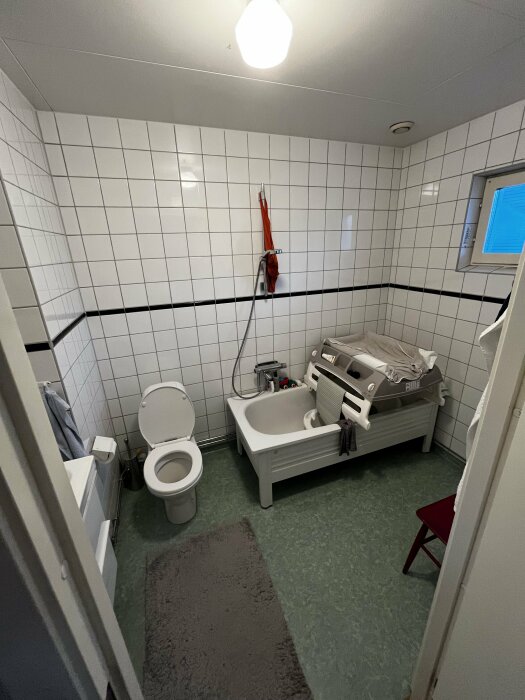 Badrum med vita kakelväggar, badkar, toalett, handfat, röd stol, matta och tvättmaskin.
