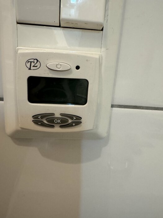 Väggmonterad enhet, troligen termostat, med display och knappar, smutsiga fläckar, T2-symbol.