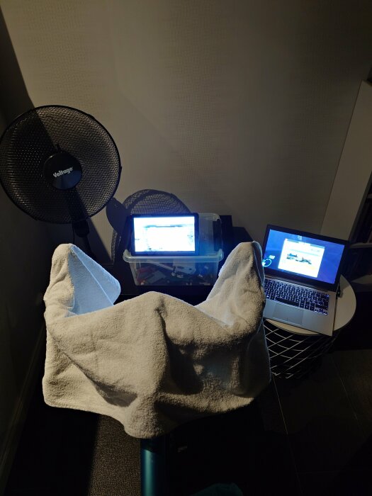 Improviserad arbetsplats med fläkt, handduk, laptop och två överlappande bord.