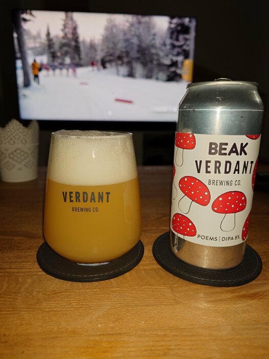 Ölglas med skum, burk från Verdant Brewing Co., skidor på TV i bakgrunden, träyta, underlägg.