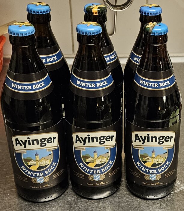 Tre flaskor Ayinger Winter Bock öl, mörka glasflaskor med blå etiketter och kapsyler.