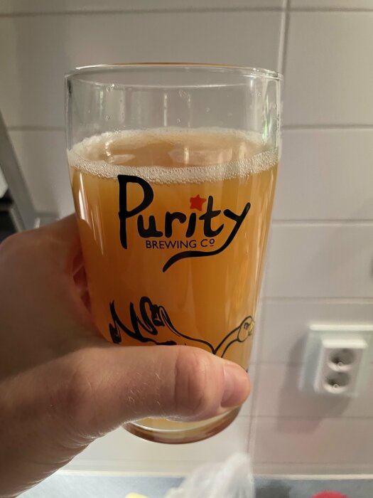 Ett glas öl hålls upp, Purity Brewing Co-logotyp, gulbrun vätska, skumtopp, inomhusmiljö, vit kaklad vägg.