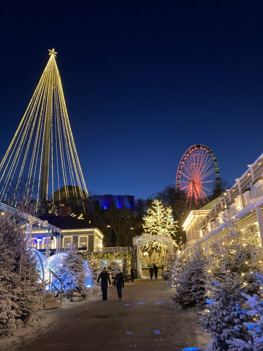 Vinterparker med upplyst julgran, pariserhjul, snö, stjärna, och promenerande människor i skymningen.