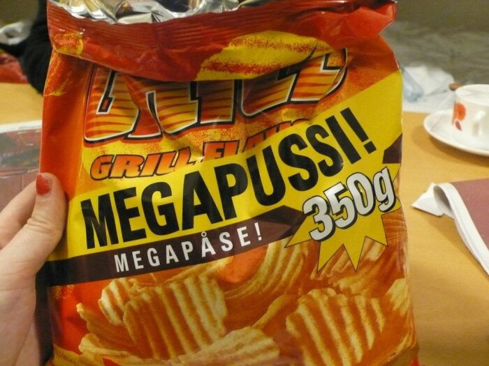 En hand håller en påse grillflavored chips märkt "MEGAPUSS", 350 gram, mot en suddig bakgrund.