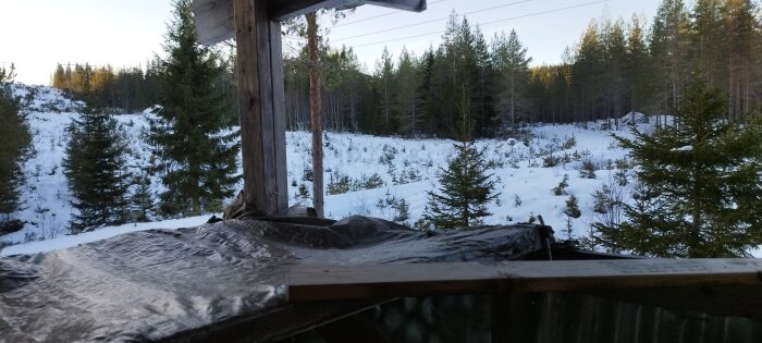 Snett trädäck, täckt i plast, med utsikt över ett snöigt skogslandskap och kyligt ljus.